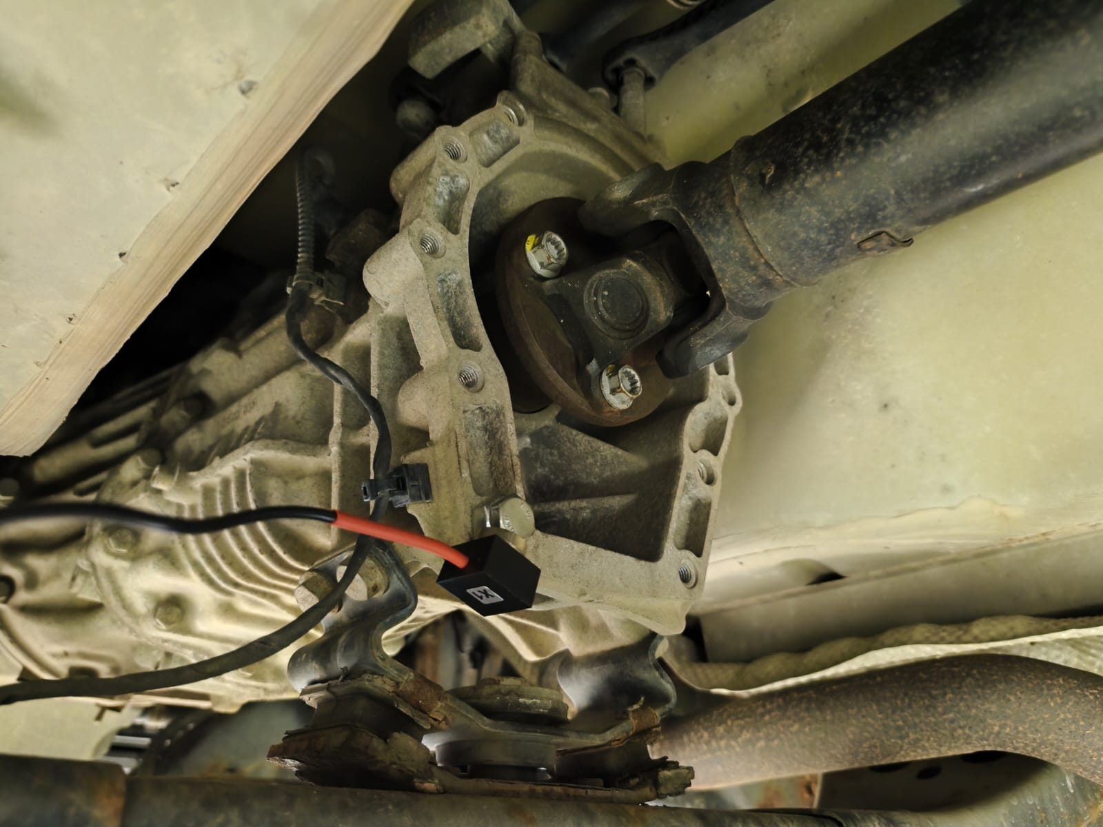 Balanset vibration sensor clamped to the rear final drive housing next to the driveshaft flange, measuring vibration on the car without removing the driveshaft