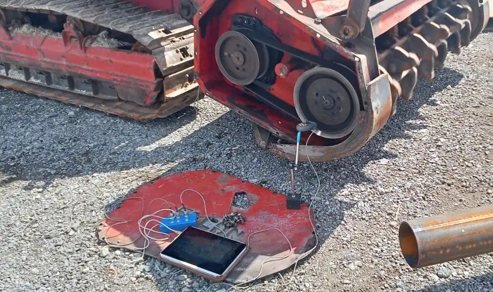 On-site balancing of a tracked mulcher rotor with Balanset-1A: measuring unit, tablet and sensors set up on the ground next to the machine